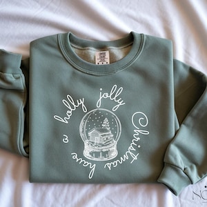 May include: Sage green sweatshirt with the words "holly jolly Christmas" and a snow globe graphic in white. The snow globe features a house and a Christmas tree. The sweatshirt has a crew neck and long sleeves.