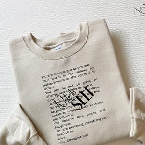 May include: A beige sweatshirt with the text "You are enough, just as you are" and other inspirational text in black. The sweatshirt has a crew neck and long sleeves. The brand name "NØA" is in the top right corner.