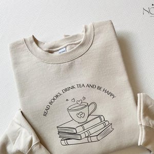May include: A beige sweatshirt with the words "READ BOOKS, DRINK TEA AND BE HAPPY" printed in black. The design shows a teacup with hearts above a stack of books. The sweatshirt is folded.