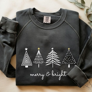 Könnte beinhalten: Dunkelgraues Sweatshirt mit Rundhalsausschnitt und langen Ärmeln. Auf der Vorderseite befinden sich vier weiße Weihnachtsbäume mit goldenen und weißen Sternakzenten. Die Worte "merry & bright" sind darunter in Schreibschrift geschrieben.