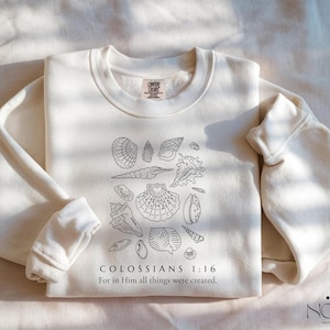 May include: Cream-colored sweatshirt featuring a black line drawing of various seashells. The text "COLOSSIANS 1:16 For in Him all things were created" is printed below the seashells. The sweatshirt has a crew neck and long sleeves.