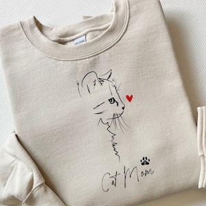 Puede incluir: Una sudadera beige con un dibujo de línea negra de la cara de un gato y el texto "Cat Mom" con una huella de pata debajo. Un corazón rojo está por encima de la cabeza del gato.