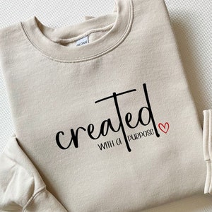 Könnte beinhalten: Ein beiger Pullover mit dem Text "created with a purpose" in schwarzer Schrift und einem kleinen roten Herzen.