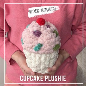 May include: A soft, plush cupcake in pink, white, and other colours. The cupcake has a textured look, with a red pom-pom on top and colourful accents. The words "VIDEO TUTORIAL" and "CUPCAKE PLUSHIE" are displayed.