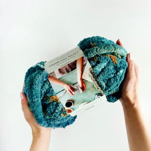 Op de afbeelding: Een bol chenille garen in de kleuren turquoise, groen en goud. Het garen is zacht en pluche, perfect voor het maken van dekens, sjaals en andere knusse items. Het label vermeldt "Chenille-Garn SELECT" en toont een foto van een vrouw die met het garen breit.