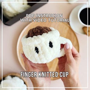Puede incluir: Una taza tejida con los dedos, de color crema con detalles negros, rellena de hilo marrón, sostenida por una persona. El texto "PDF Instruction with Video Tutorial" y "Finger Knitted Cup" son visibles.