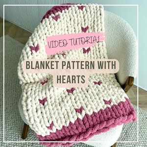 Könnte beinhalten: Eine weiße und rosafarbene Strickdecke mit einem Herzmuster. Die Decke liegt über einem Stuhl und der Text "VIDEO TUTORIAL BLANKET PATTERN WITH HEARTS" ist sichtbar.