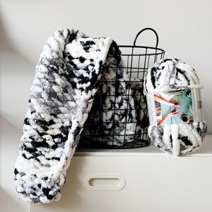 Op de afbeelding: Een zwart-wit garenklos met een label met de tekst "Extra Super Soft Chenille" in een zwarte draadmand met een zwart-wit pluche deken die over de rand hangt.