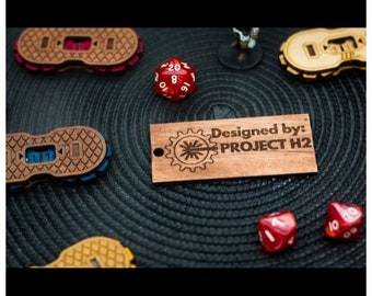 Puede incluir: Un letrero de madera con el texto "Designed by: PROJECT H2" y un diseño de engranaje. El letrero está sobre una superficie negra con dados rojos y amarillos.