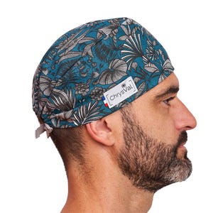 Op de afbeelding: Turquoise en grijze chirurgische scrubcap met bloemenpatroon en de tekst "ChryVal" op een wit label. De cap heeft een stropdas aan de achterkant voor een verstelbare pasvorm. Ontworpen voor medisch professionals.