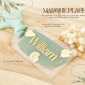 Peut inclure: Une étiquette de place en acrylique transparent avec le nom "William" en lettres dorées, sur fond vert sauge. Des accents de feuilles d'or et un ruban vert menthe ajoutent des détails. Le texte dit "MARQUE PLACE".