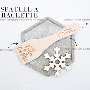 Peut inclure: Une spatule en bois avec le texte "C'EST le Roi DE LA Raclette" et le nom "CYRIL" gravé dessus. La spatule est sur une surface hexagonale grise avec un flocon de neige blanc.