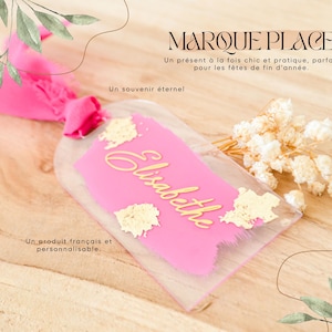 Op de afbeelding: Een heldere acryl naamkaart met een roze middenstuk en gouden letters met de tekst "Elisabeth". De kaart is vastgebonden met een roze lint en versierd met bladgoud accenten en gedroogde bloemen. Tekst op de kaart luidt "MARQUE PLACE."