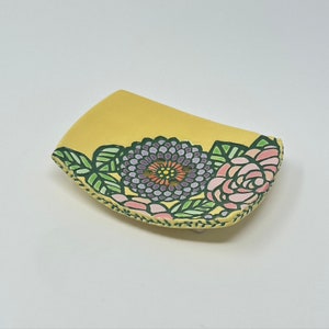 May include: A yellow ceramic soap dish with a floral design. The dish features a large purple flower with a yellow center and pink and white flowers with green leaves.