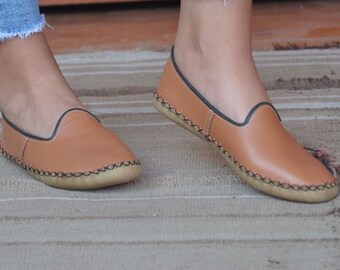 May include: A pair of brown leather slip-on shoes with black stitching around the edges. The shoes have a flat sole and are worn by a person wearing blue jeans.