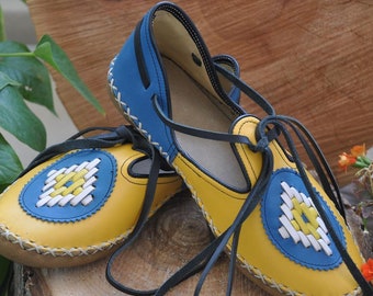 May include: A pair of yellow leather sandals with blue and white embroidered details. The sandals have black laces that tie around the ankles.