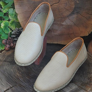 May include: A pair of beige slip-on shoes with a perforated pattern and brown trim. The shoes are sitting on a wooden stump.