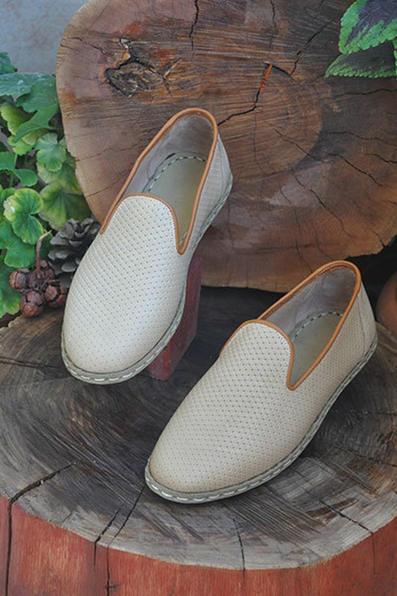 May include: A pair of beige slip-on shoes with a perforated pattern and brown trim. The shoes are sitting on a wooden stump.