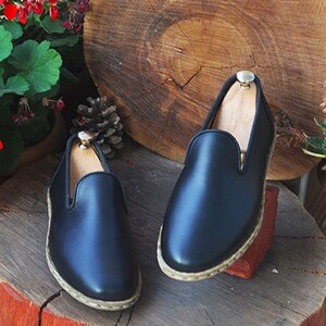 May include: Two pairs of black leather slip-on shoes with a woven trim around the sole. The shoes are sitting on a wooden stump.