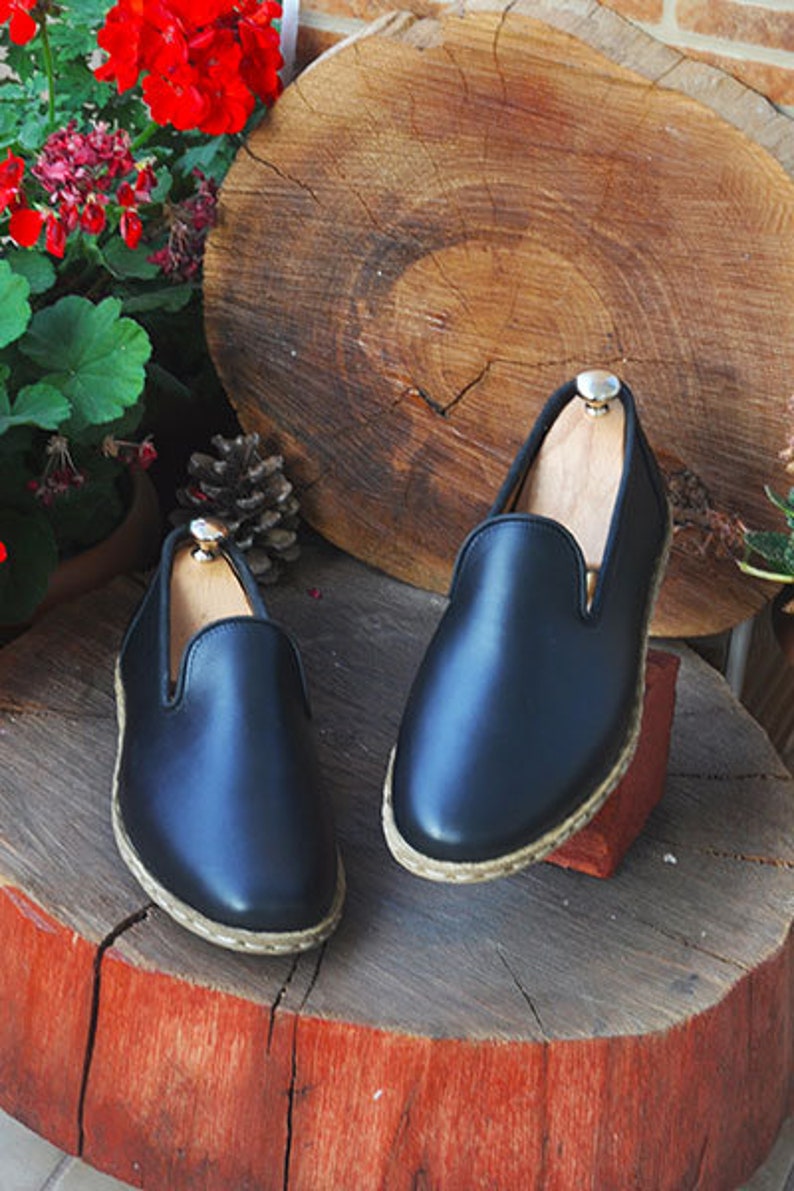 May include: Two pairs of black leather slip-on shoes with a woven trim around the sole. The shoes are sitting on a wooden stump.