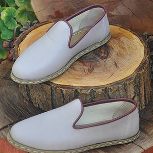 May include: Two pairs of light gray leather slip-on shoes with a burgundy trim around the top edge. The shoes have a white stitched edge around the sole.