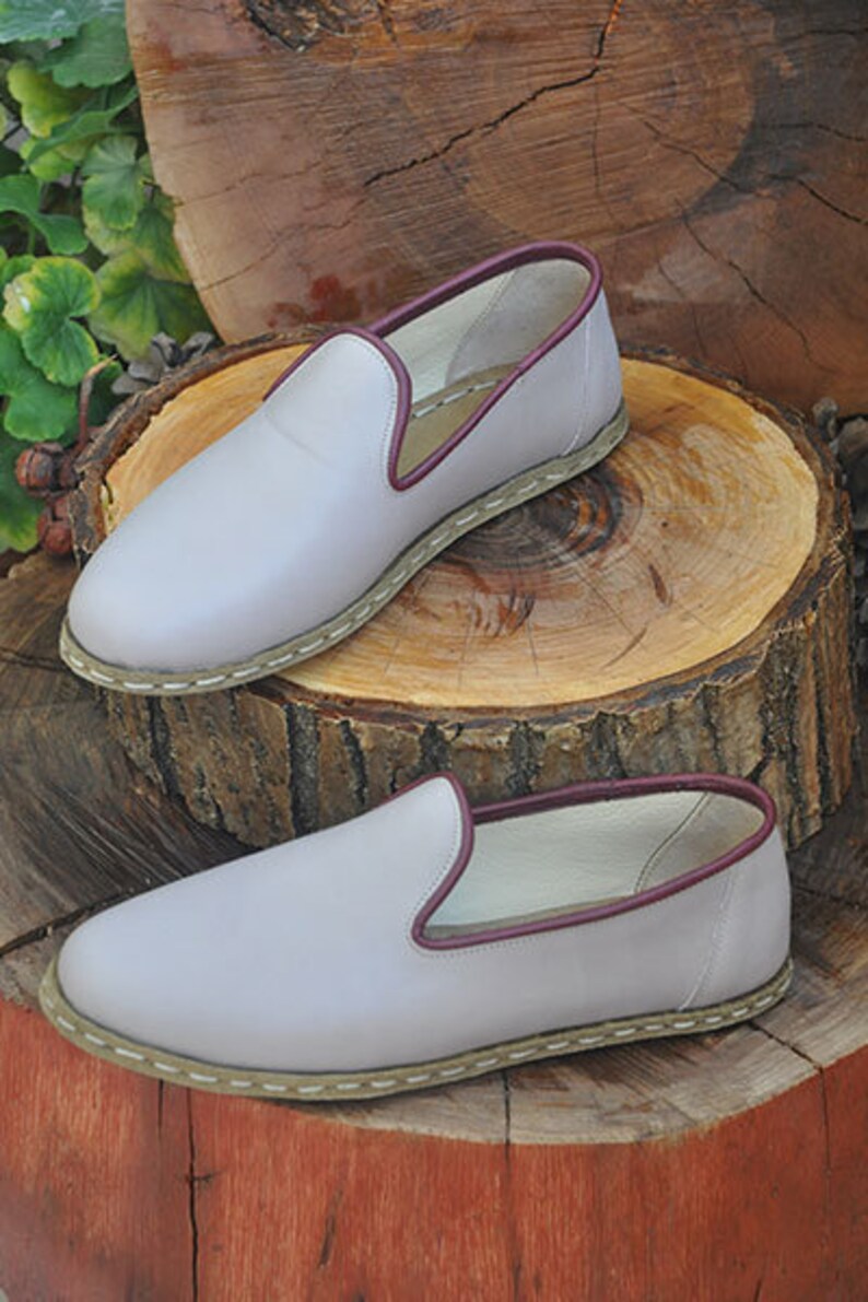 May include: Two pairs of light gray leather slip-on shoes with a burgundy trim around the top edge. The shoes have a white stitched edge around the sole.