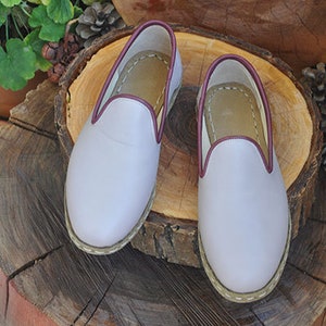 May include: A pair of light purple leather slippers with a burgundy trim. The slippers are sitting on a wooden surface.