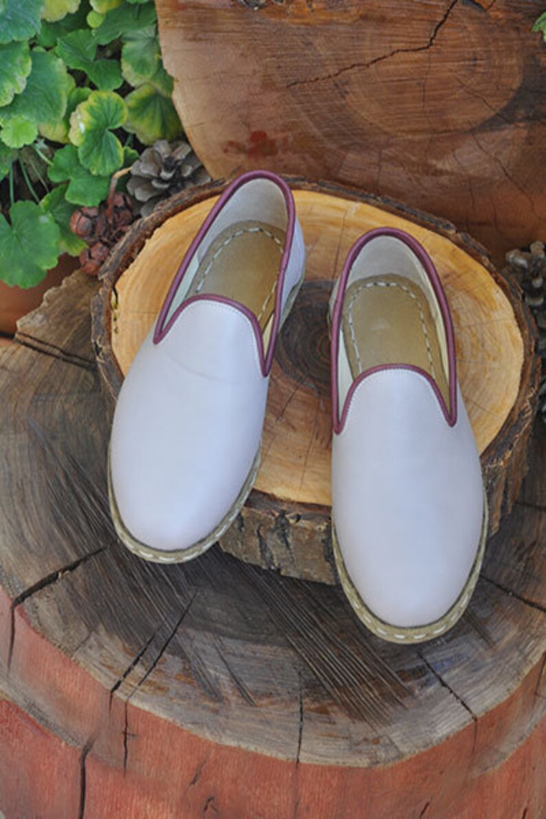 May include: A pair of light purple leather slippers with a burgundy trim. The slippers are sitting on a wooden surface.