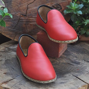 May include: Two red leather slip-on shoes with black trim and a white stitching detail around the sole. The shoes are sitting on a wooden stump.