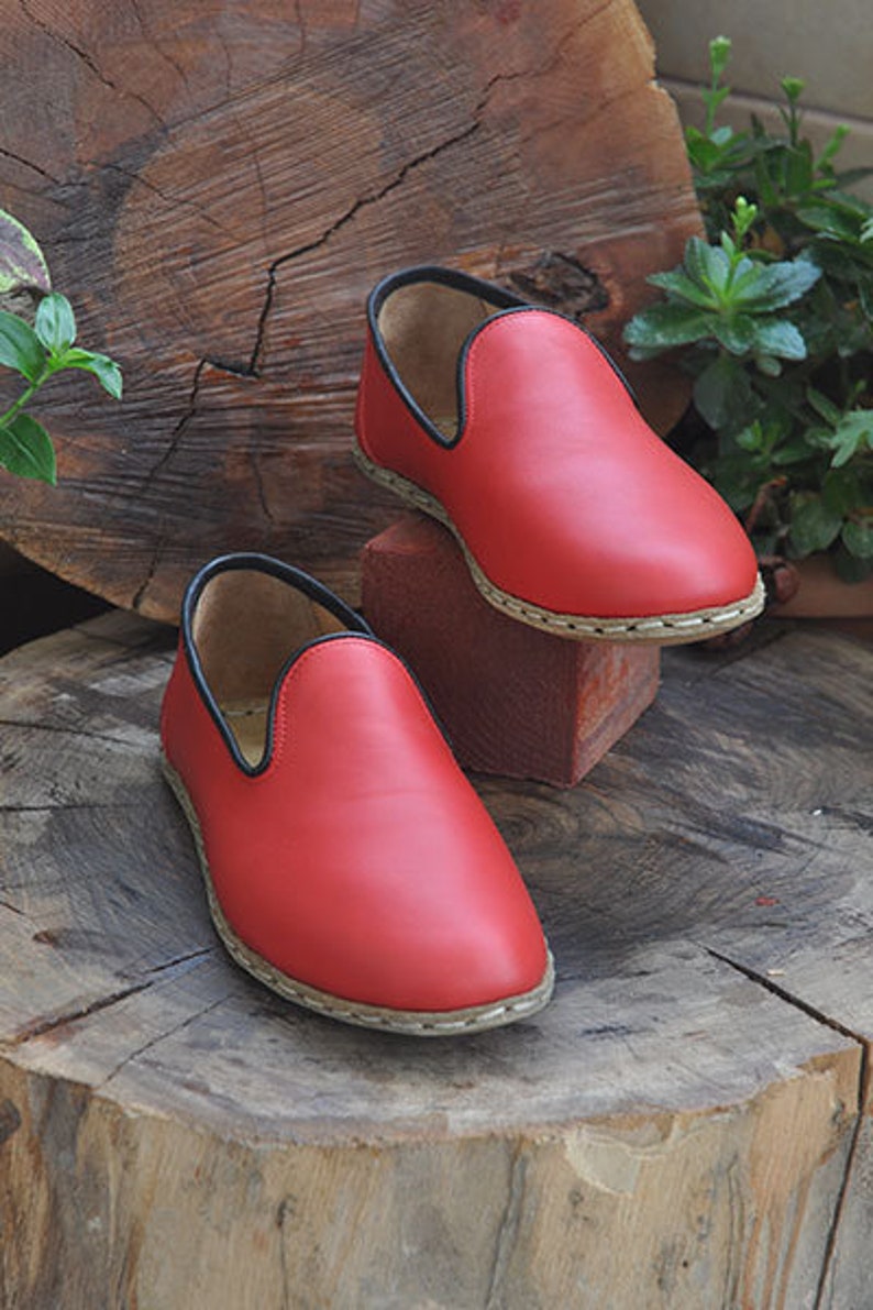 May include: Two red leather slip-on shoes with black trim and a white stitching detail around the sole. The shoes are sitting on a wooden stump.