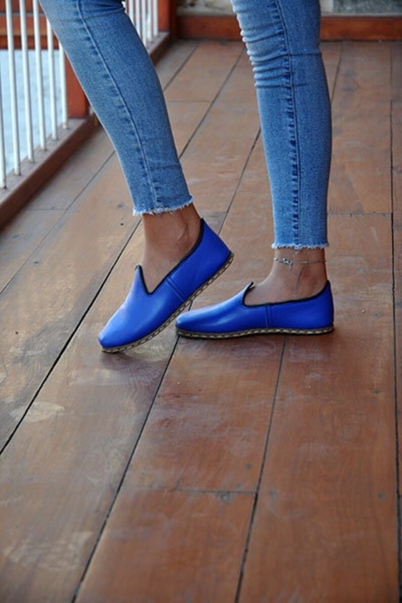 May include: A pair of blue leather slip-on shoes with a black trim. The shoes are worn by a person wearing blue jeans.