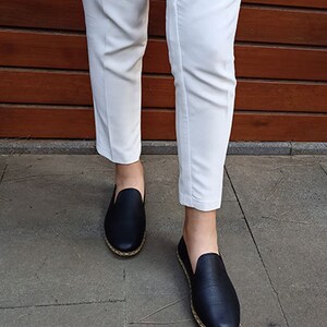 May include: A pair of black leather slip-on shoes with a gold trim around the sole. The shoes are being worn by a person wearing white pants.