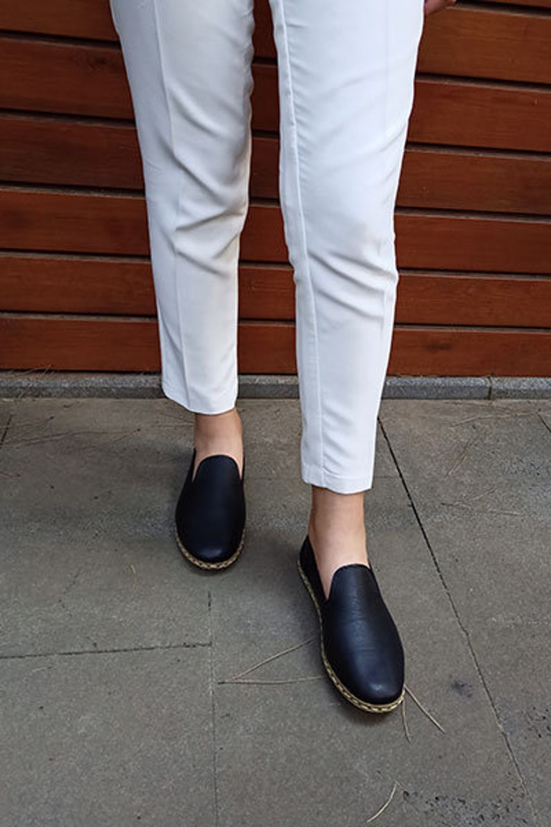 May include: A pair of black leather slip-on shoes with a gold trim around the sole. The shoes are being worn by a person wearing white pants.