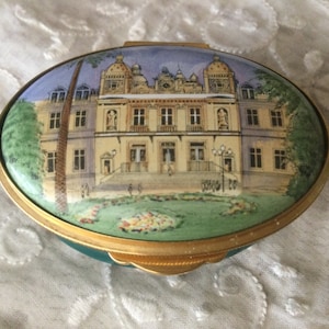 May include: Oval trinket box with a green base and a gold rim. The lid features a painted image of a large building with a light blue sky and green grass.