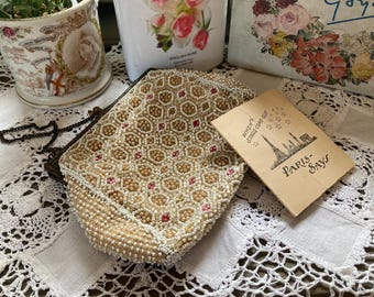 May include: A cream and gold beaded vintage handbag with a geometric design, featuring small pink beads. A card with the Eiffel Tower and the text "Paris Says" is placed next to the bag. The bag has a dark metal frame and a beaded strap.
