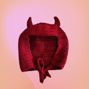 May include: A red crocheted devil hat with horns and a tie at the bottom.
