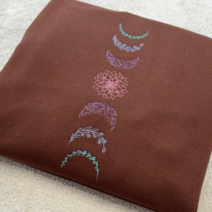 May include: A brown sweatshirt with an embroidered design of a crescent moon with floral accents in shades of pink, purple, and green.
