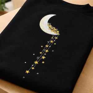 May include: Black sweatshirt with an embroidered design of a crescent moon and falling stars in yellow and white. The celestial design is on the upper left. The sweatshirt is made of a soft material. The design is reminiscent of a night sky.