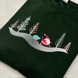 May include: A dark green sweatshirt featuring an embroidered design of a silver winding path through a forest of white pine trees with red berries. Santa Claus is depicted running along the path carrying a light blue sack over one shoulder.