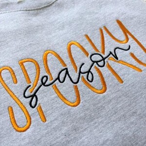 May include: A light grey sweatshirt featuring embroidered text. The word Spooky is stitched in large, bold orange letters, while the word season is stitched in a smaller, cursive black font that overlays the orange text across the centre.
