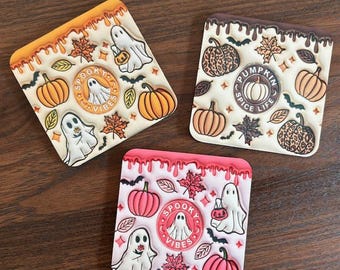 May include: Three square coasters with a Halloween theme. The coasters feature a white background with orange, brown, and pink accents. The first coaster has a dripping orange glaze and features a ghost with the text "Spooky Vibes". The second coaster has a dripping brown glaze and features a pumpkin with the text "Pumpkin Spice Life". The third coaster has a dripping pink glaze and features a ghost with the text "Spooky Vibes".