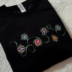 May include: Black sweatshirt featuring an embroidered design of festive Christmas lights. The lights are in jewel tones of blue, purple, red, and orange, with gold and green detailing. The design is on the lower left of the sweatshirt.
