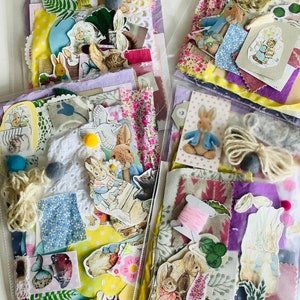 May include: A collection of fabric scraps, embellishments, and paper ephemera featuring a Peter Rabbit theme. The collection includes a variety of colors and textures, including pink, purple, blue, yellow, and green. There are also several small, white pom-poms and a few spools of thread.