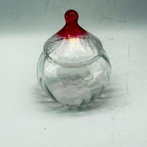 May include: A clear glass jar with a lid. The lid has a red top and a clear base. The jar has a round shape with a swirl design. The jar is empty and sits on a white surface. The jar is likely used for storage or decoration.