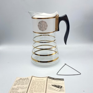 May include: A white glass coffee pot with a gold trim and a black handle. The pot has a white lid with a gold design. The pot is sitting on a white surface. The image also includes a black and white instruction sheet with the text "Handy Order Form" and "Guarantee".