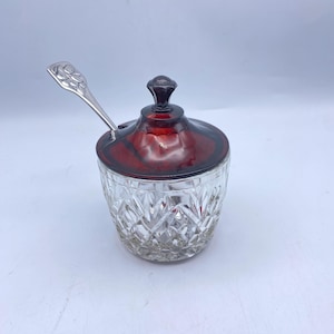 May include: A clear glass jar with a cut glass pattern and a red glass lid. The jar has a small silver spoon with a decorative handle.