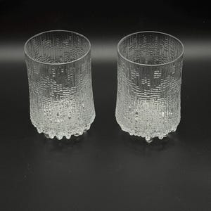 May include: Two clear, textured glass tumblers. The glasses have a cylindrical shape with a textured pattern on the sides and a decorative base. The glasses are made of clear glass and are set against a black background.