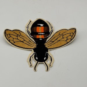 May include: An embroidered bee patch featuring a black head and thorax, with a segmented abdomen displaying horizontal stripes of black, brown, and orange. The insect has two large, textured golden-brown wings with black outlines and thin white legs.