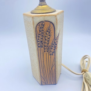 May include: A vintage ceramic table lamp with a wheat stalk design. The lamp has a beige, hexagonal base with a brown wheat stalk illustration. The lamp's metal components are gold-toned, and the electrical cord is beige.