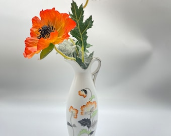 May include: A white ceramic vase with a floral design and a silver rim. The vase is filled with two orange poppy flowers.